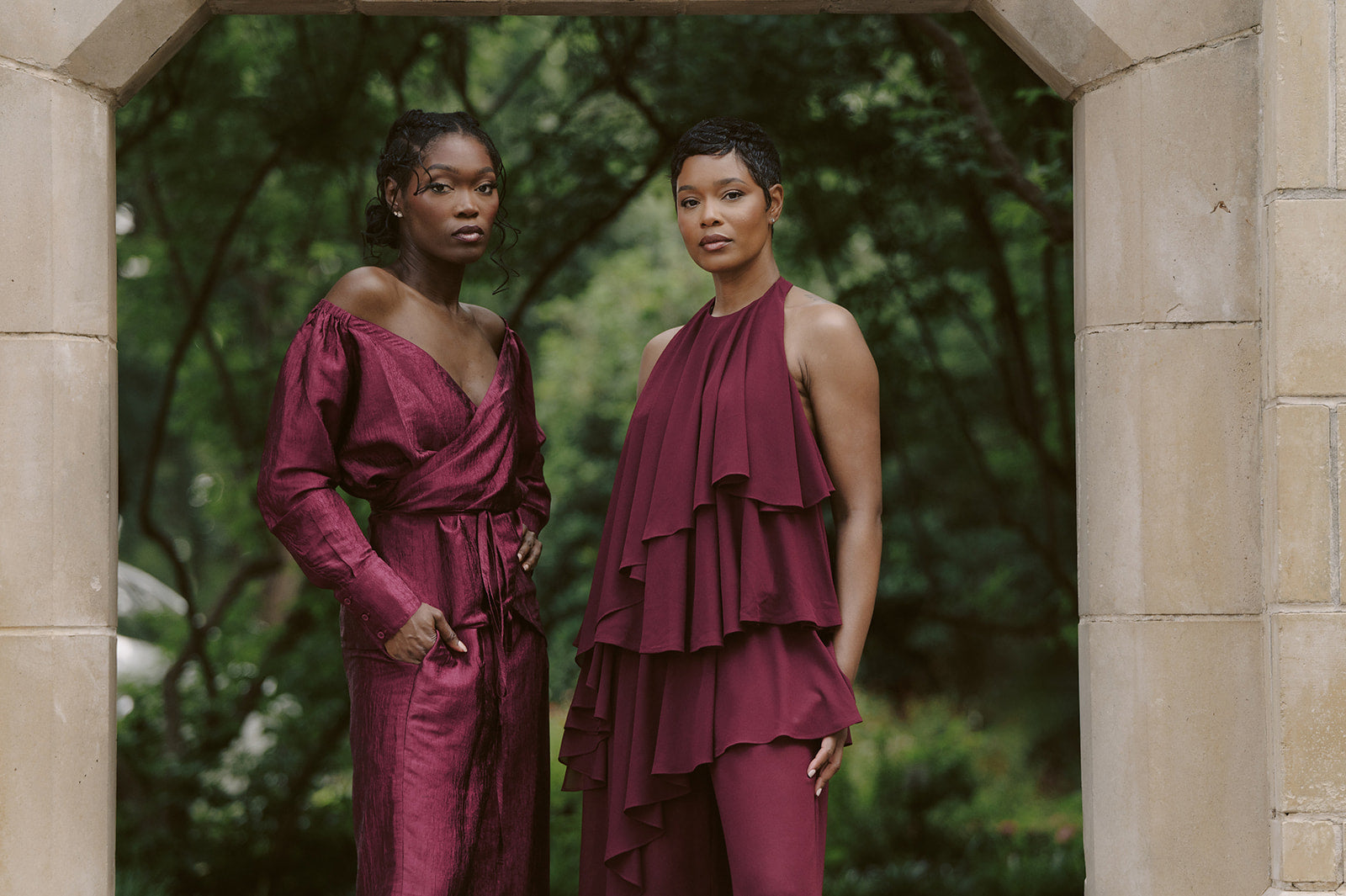 Two tall women in burgundy dresses standing in a stone archway with greenery behind them.